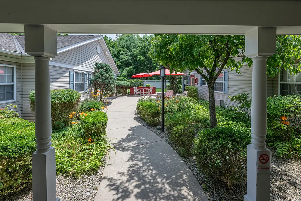 Sequence (1)Cedar Ridge-19 A paved walkway between garden areas leads to a patio with red umbrellas and outdoor seating, surrounded by residential buildings.