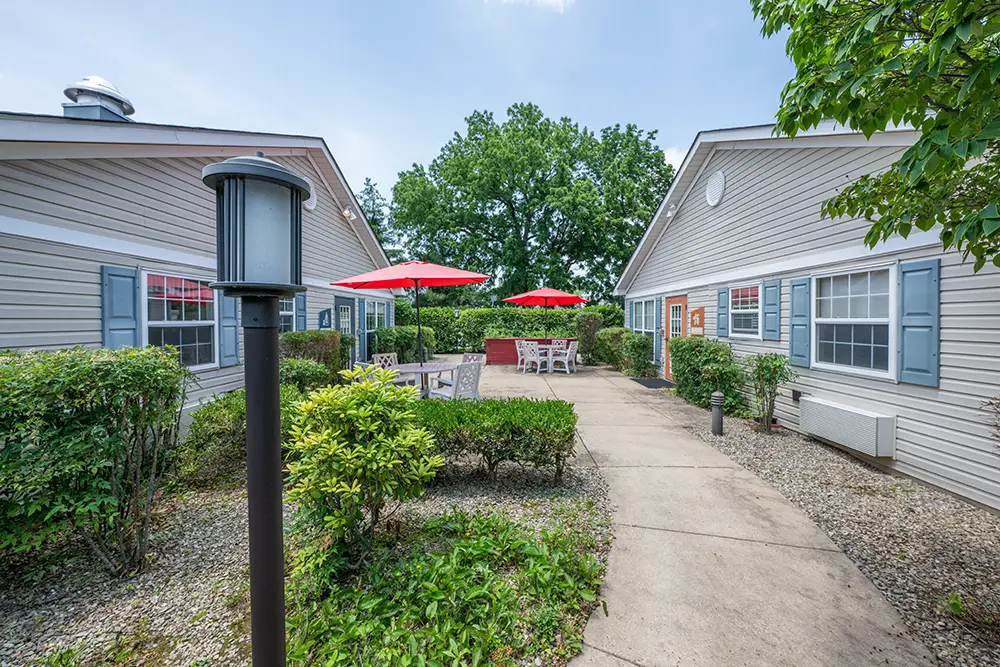 Sequence (1)Cedar Ridge-2 A paved walkway leads between two single-story buildings to an outdoor patio area with tables, chairs, and red umbrellas surrounded by greenery.