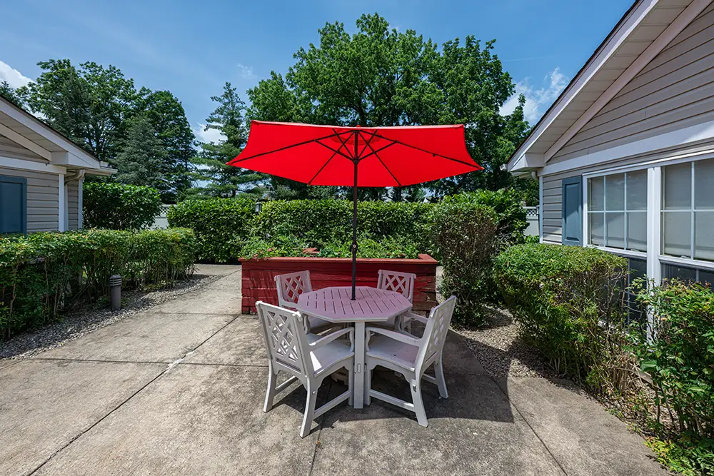 Sequence (1)Cedar Ridge-5 A round patio table with four white chairs and a large red umbrella sits on a concrete patio between two buildings, surrounded by green bushes and trees.