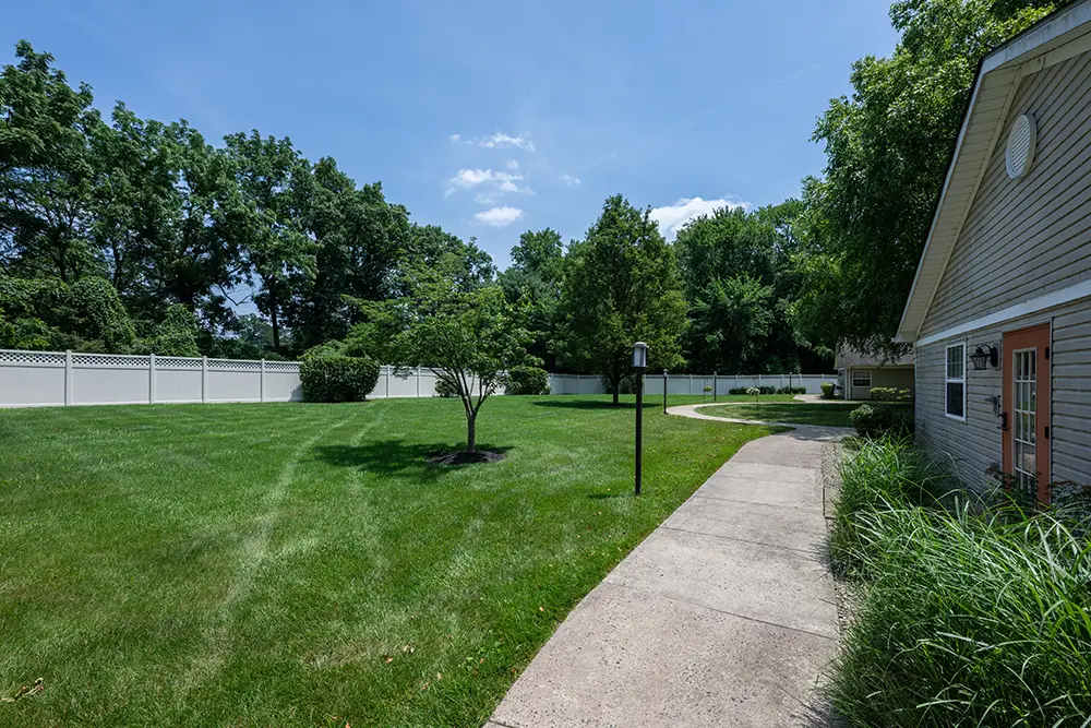 Sequence (1)Cedar Ridge-6 A concrete walkway curves through a green lawn with a small tree, bordered by a white fence and trees, next to a building with beige siding and a red door.