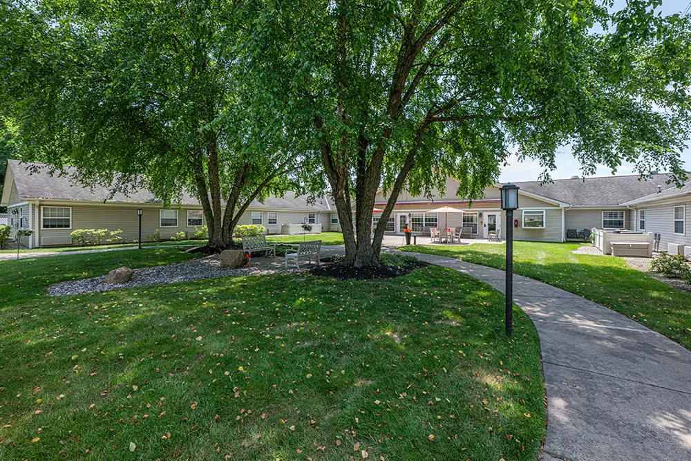 Sequence (1)Cedar Ridge-7 Curved concrete walkway and outdoor seating area shaded by large trees in the landscaped courtyard of a residential building.