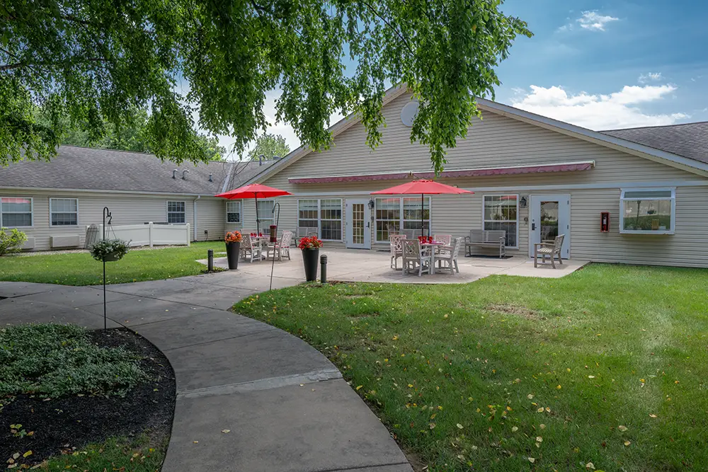 Sequence (1)Cedar Ridge-8 A paved walkway leads to a patio with tables, chairs, and red umbrellas outside a beige building, surrounded by grass and trees under a partly cloudy sky.