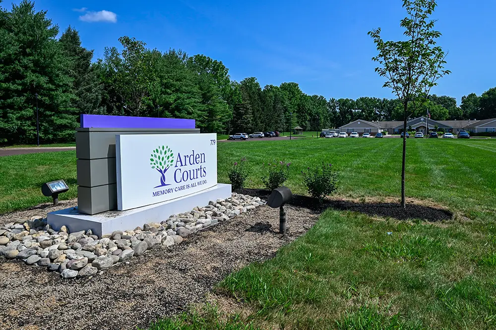 Sequence (1)Cedar Ridge signs A stone sign reads "Arden Courts Memory Care & AL/IL" on a grassy lawn with trees, a small tree, and a distant building in the background under a blue sky.