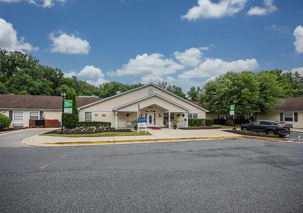 Single-story building with a gabled roof, landscaped front yard, and a driveway in front, Arden Courts specializes in memory care, set against a backdrop of trees and a partly cloudy Silver Spring sky.