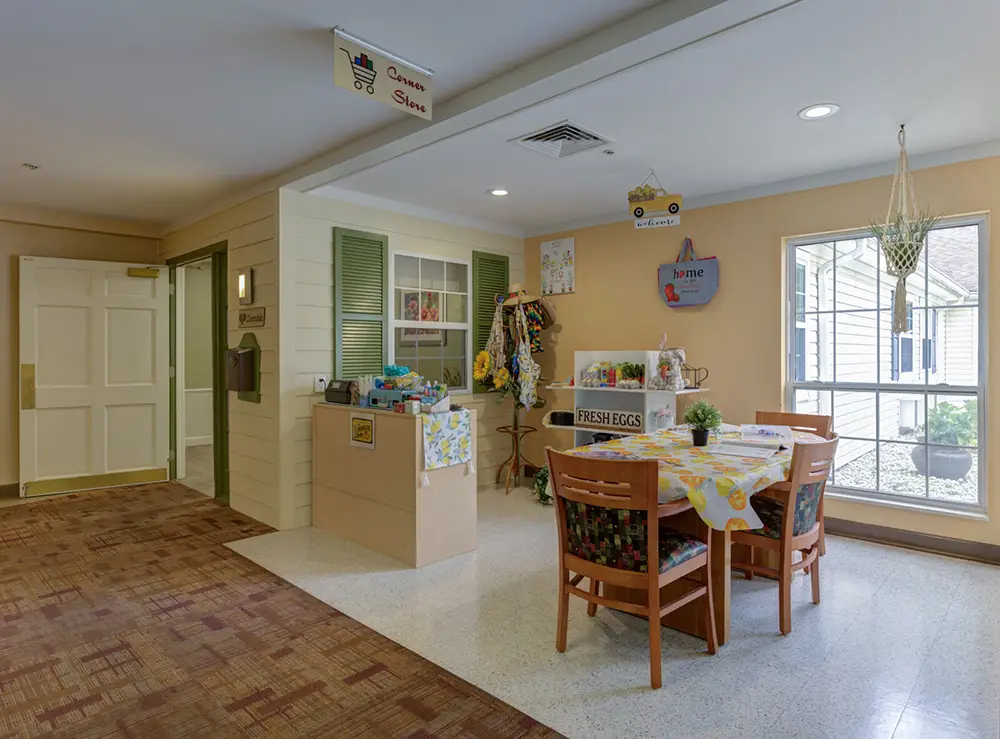 A small indoor shop area with a display counter, a round table with four chairs, and various decorations, next to a large window and double doors.