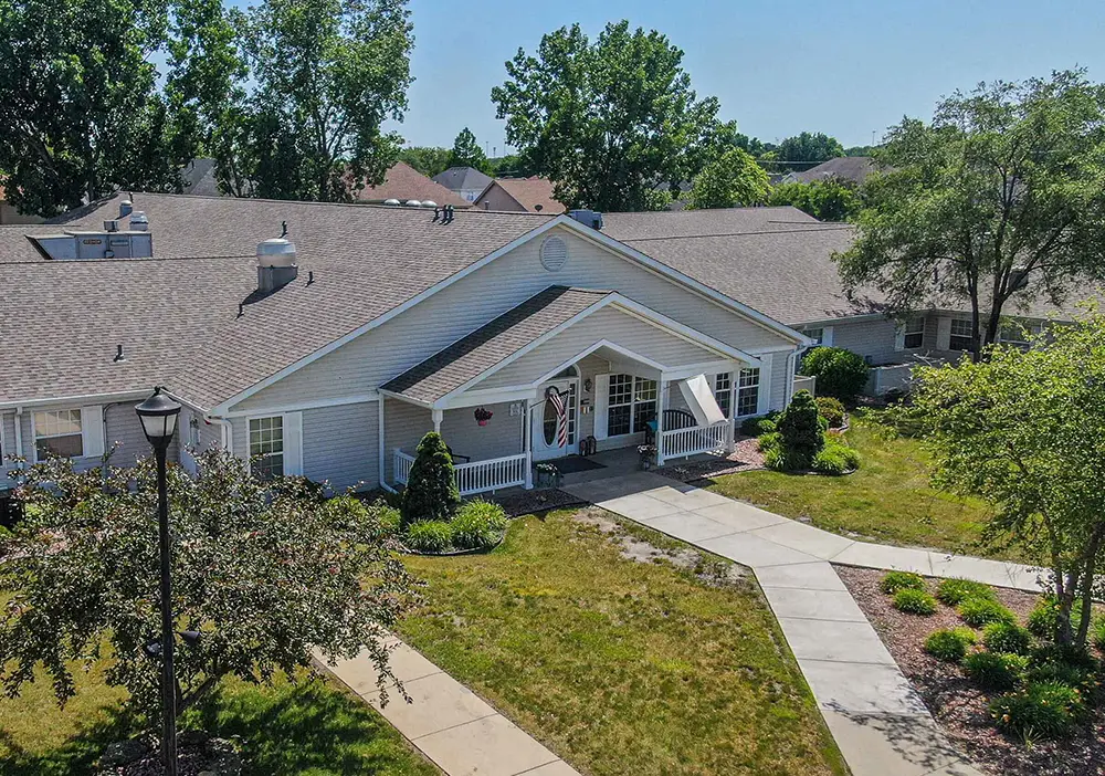 A single-story residential building in South Holland with a covered porch, surrounded by trees, shrubs, and pathways on a sunny day—perfect for memory care at Arden Courts.