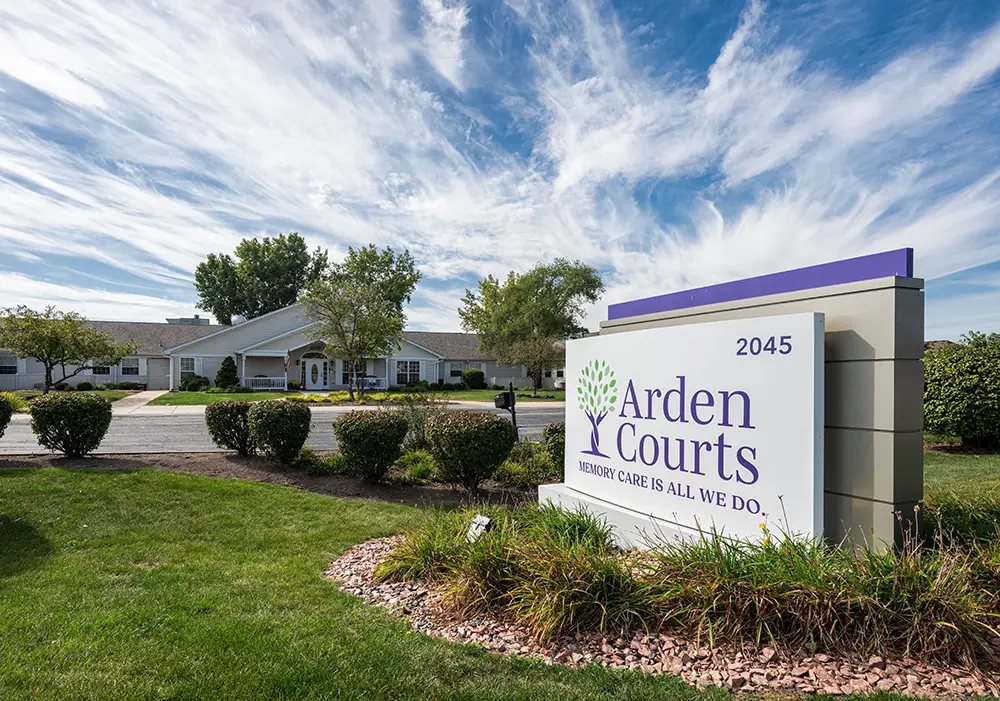 South Holland New photos04 A white building surrounded by greenery with a large sign in front reading "Arden Courts, Memory Care is All We Do, 2045" under a blue sky with wispy clouds.