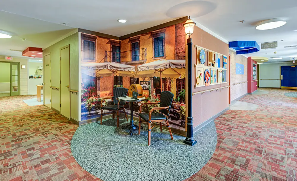 Indoor hallway with a mural of a European café scene, two chairs and a table, a streetlamp, and artwork displayed on the wall.
