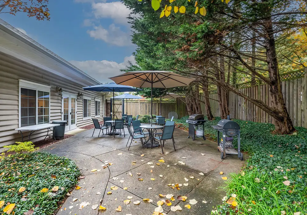 Outdoor patio area at Springhouse Senior Living with tables, chairs, two umbrellas, two grills, and a fenced yard surrounded by trees and greenery. Fallen leaves are scattered on the concrete surface, creating a peaceful senior living retreat.