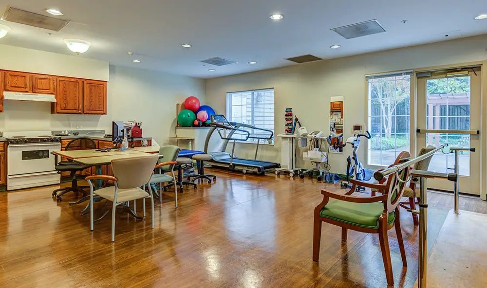 A rehabilitation room with exercise equipment, therapy balls, a treadmill, chairs, tables, and large windows letting in natural light. A rehabilitation room with exercise equipment, therapy balls, a treadmill, chairs, tables, and large windows letting in natural light.