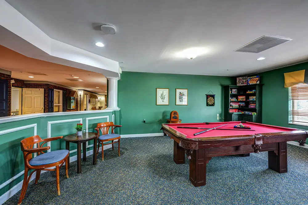 Room with a green accent wall, pool table, cue sticks, dartboard, bookshelf with board games, and a small table with two chairs.