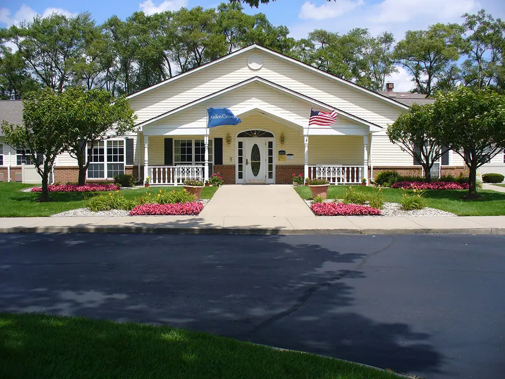 Single-story building with a white front porch, flower beds, and two flags on display; Arden Courts in Sterling Heights offers memory care surrounded by green lawns and trees under a clear sky.