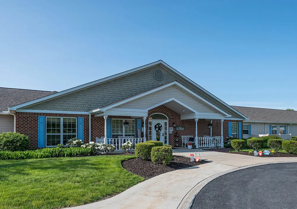 Single-story brick building with white columns, blue shutters, manicured bushes, and a curved driveway welcomes you to Arden Courts of Susquehanna, offering specialized memory care under a clear blue sky.