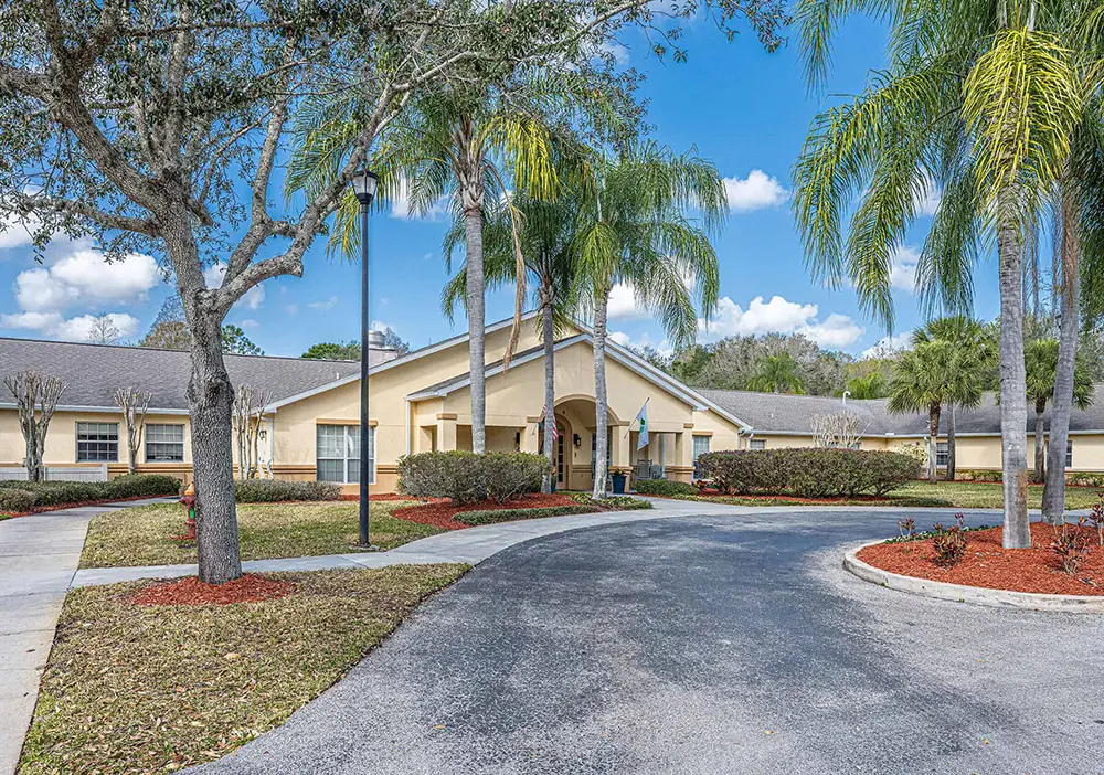 Single-story beige building with a gabled roof, palm trees, a curved driveway, and neatly landscaped grounds under a blue sky—Arden Courts in Tampa specializes in memory care for seniors.