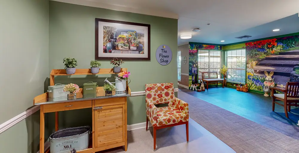 A small flower shop-themed area with potted plants, a floral chair, and wall art in a brightly lit hallway with colorful mural and windows.