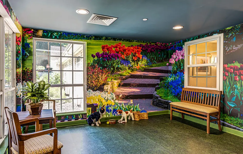 Room with a garden mural on the wall, wooden benches, a chair, a potted plant, toy dog figures, and windows letting in natural light.