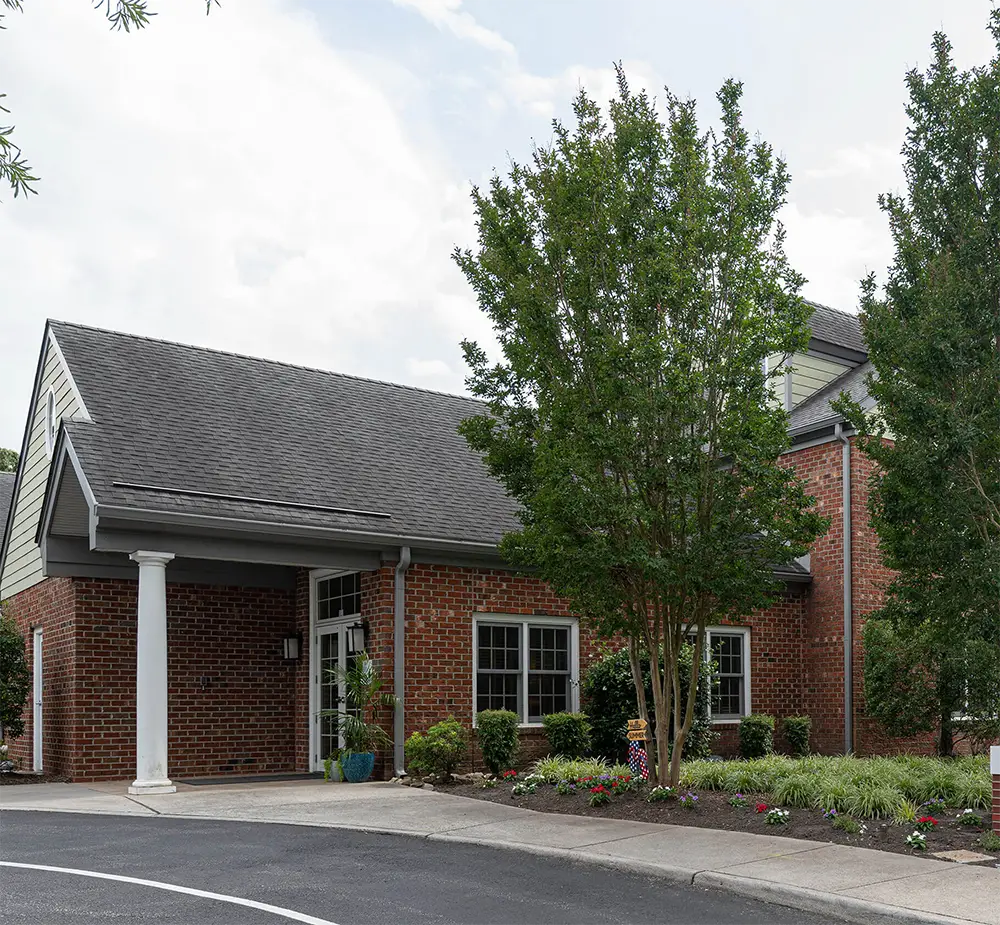 Arden Courts in Virginia Beach is a brick building with white columns, large windows, and landscaped greenery at the entrance, seen from a curved driveway on a cloudy day, offering specialized memory care services.