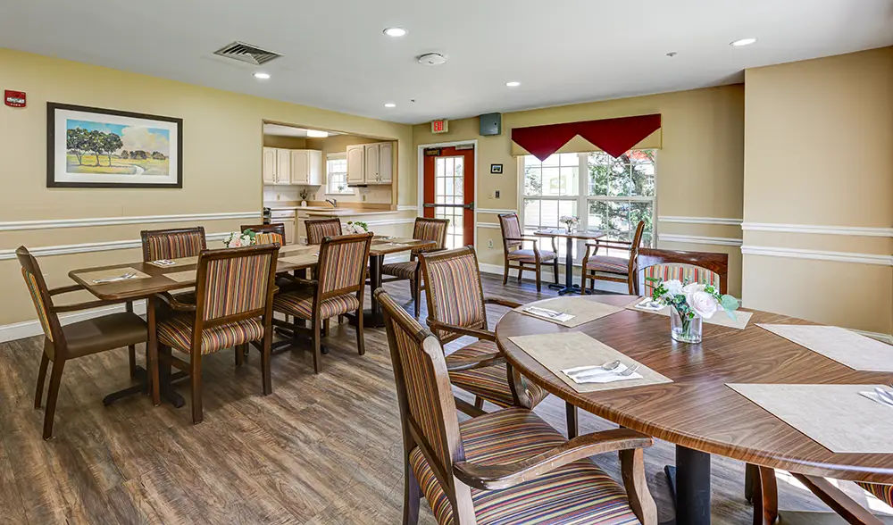 WAR_9078_79_80_81_82 A dining area with several wooden tables and striped chairs, placemats on the tables, floral centerpieces, and a view into a kitchen with natural light from windows.