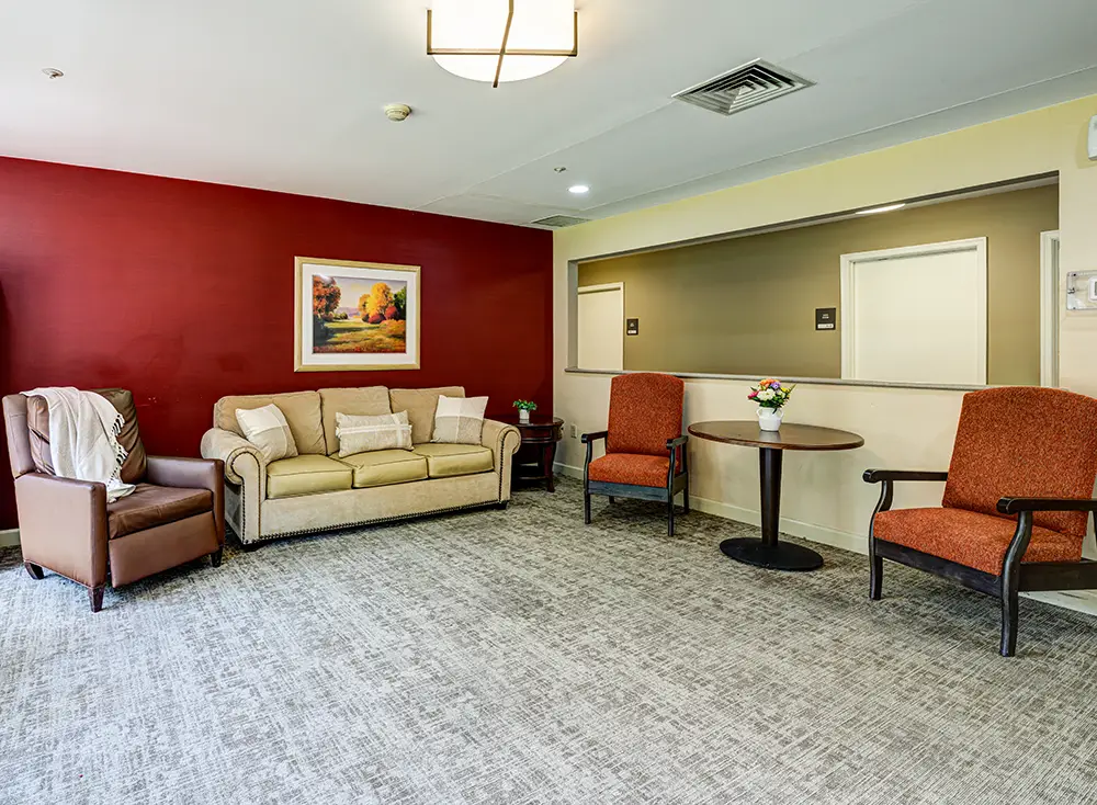 WAR_9103_4_5_6_7 A waiting room with a beige sofa, two orange chairs, a round table with flowers, brown armchair, red accent wall, and framed landscape painting.