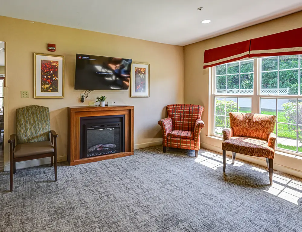 WAR_9108_09_10_11_12 A small living room with two patterned armchairs, a wooden fireplace, a TV mounted above, framed artwork, and a large window letting in natural light.