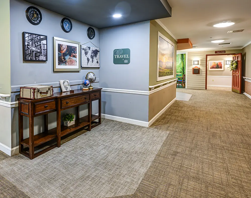 WAR_9113_4_5_6_7 A carpeted hallway features travel-themed decor, framed art, a small wooden table with a globe, and neutral wall colors with overhead lighting.