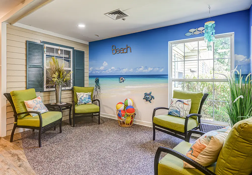WAR_9128_29_30_31_32 A waiting room features beach-themed decor, green cushioned chairs, a beach mural, a window with garden view, and a basket of colorful balls on the carpeted floor.