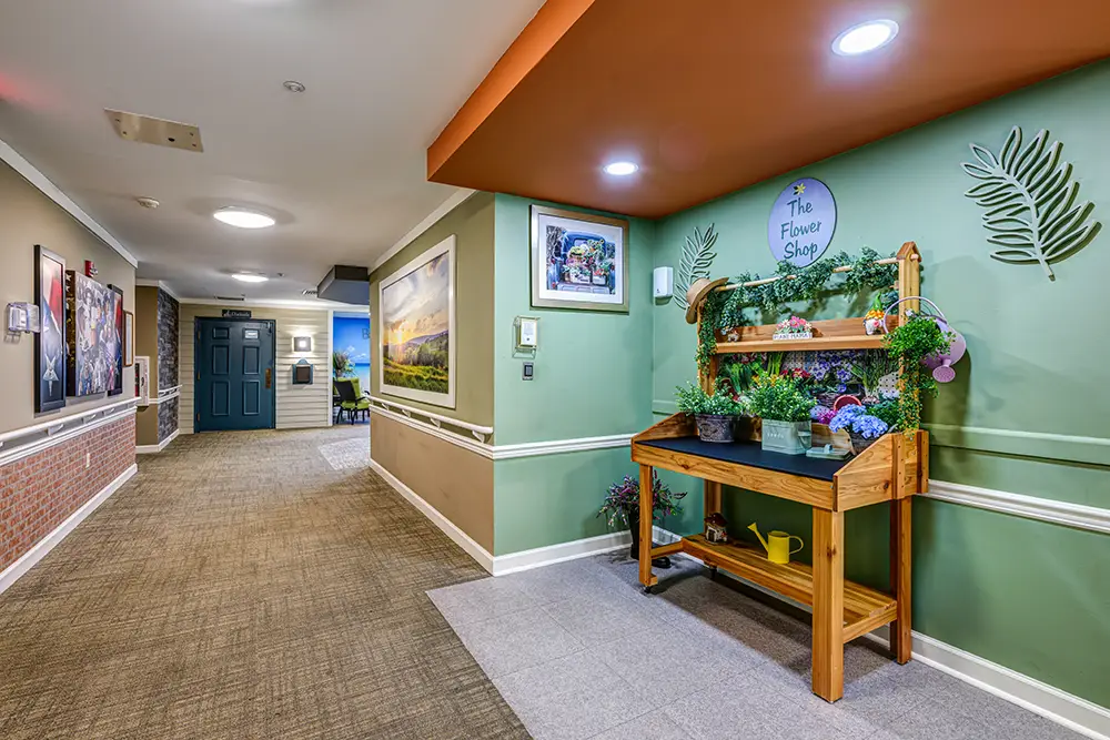 WAR_9138_39_40_41_42 A hallway with carpeted floors and framed art features a wooden flower stand labeled "The Flower Shop," decorated with plants and gardening tools beneath bright ceiling lights.