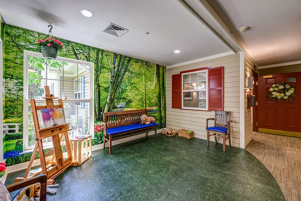 WAR_9153_4_5_6_7 A cozy indoor area with a bench, an easel, and a large wall mural of a forest scene, decorated with potted plants and a teddy bear.