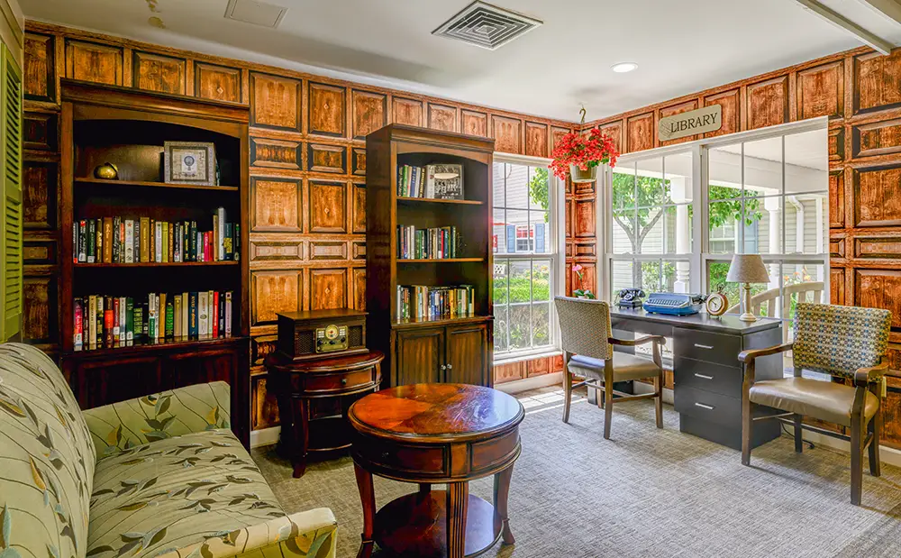 WAR_9168_69_70_71_72 A cozy library room with wooden bookshelves, a floral sofa, a round coffee table, a desk with two chairs, and large windows letting in natural light.
