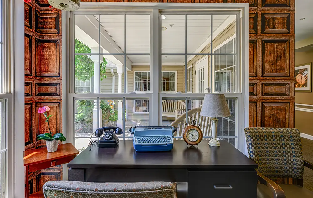 WAR_9173_4_5_6_7 A desk with a blue typewriter, rotary phone, lamp, clock, and potted plant is positioned in front of a window overlooking a porch with rocking chairs.