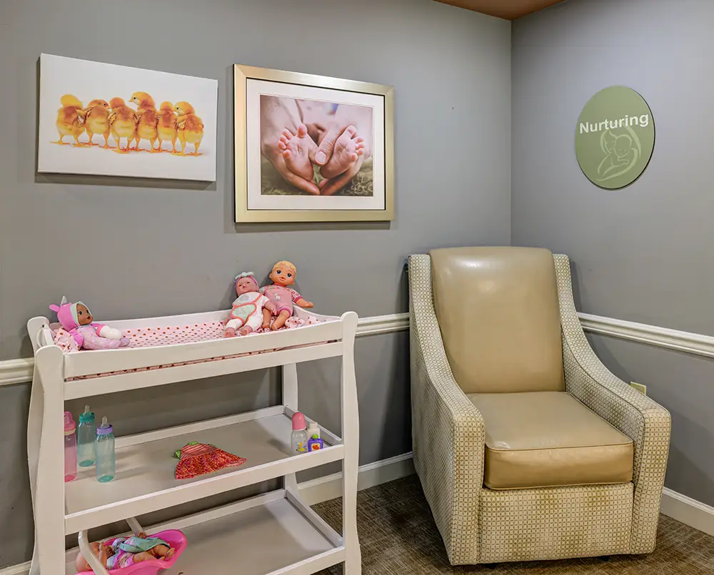 WAR_9188_89_90_91_92 A beige armchair sits next to a changing table with baby dolls, bottles, and toys. The wall has framed photos and a sign reading "Nurturing.