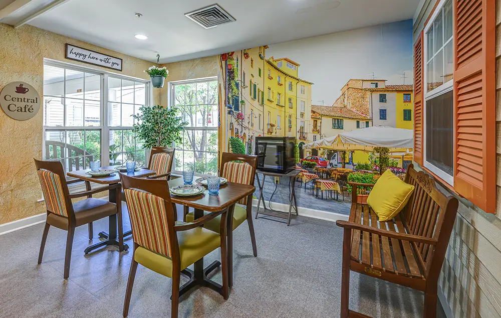 WAR_9208_09_10_11_12 A small café-style seating area with two tables, a wooden bench, and a wall mural depicting a colorful European street scene.