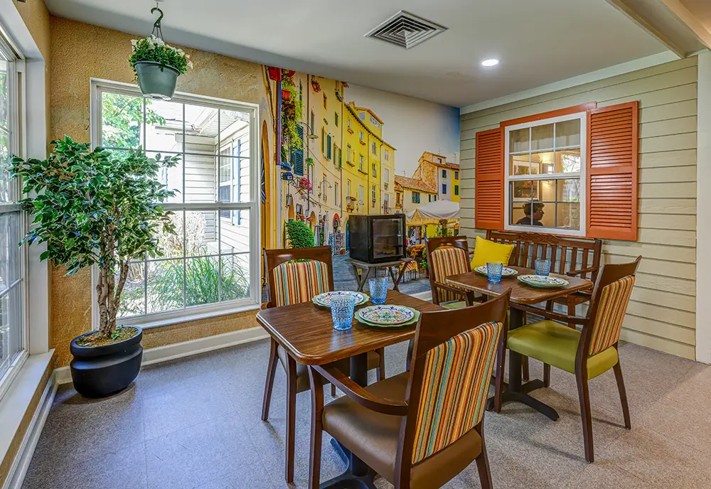 WAR_9213_4_5_6_7 A dining area with two wooden tables set for four, surrounded by chairs, indoor plants, large windows, and a mural depicting a colorful European street scene.