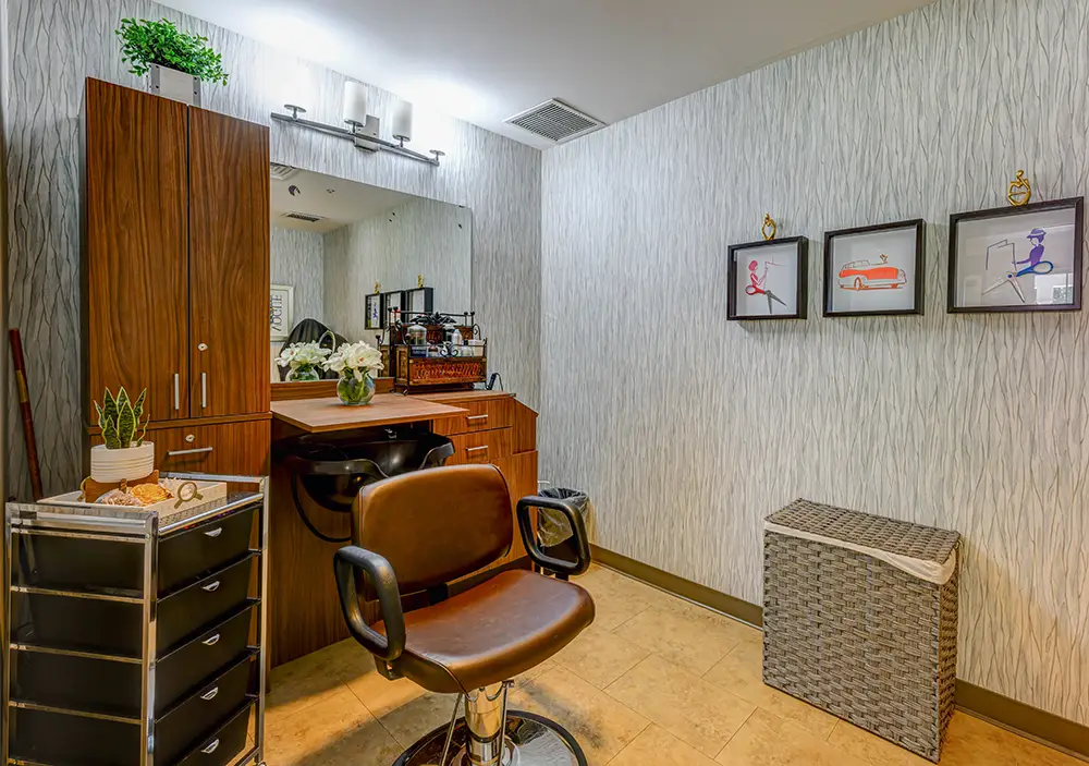 WAR_9223_4_5_6_7 A salon station with a brown chair, wooden cabinets, mirror, plants, and three framed pictures on a textured wall. A wicker hamper and storage cart are also visible.