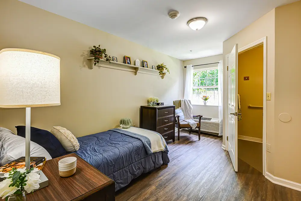 WAR_9238_39_40_41_42 A neatly arranged bedroom with a single bed, a nightstand, a dresser, a chair by the window, and a shelf with small decorations on a beige wall.