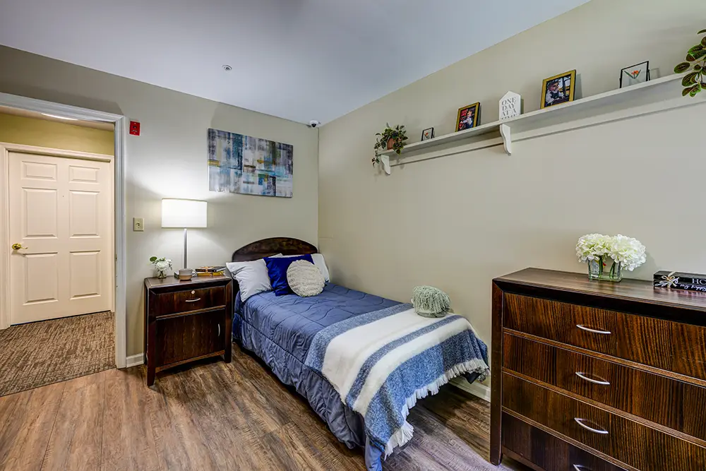 WAR_9248_49_50_51_52 A small, neatly made bed with a blue comforter is next to a nightstand and dresser in a tidy, well-lit bedroom with framed art and decorative items on a wall shelf.