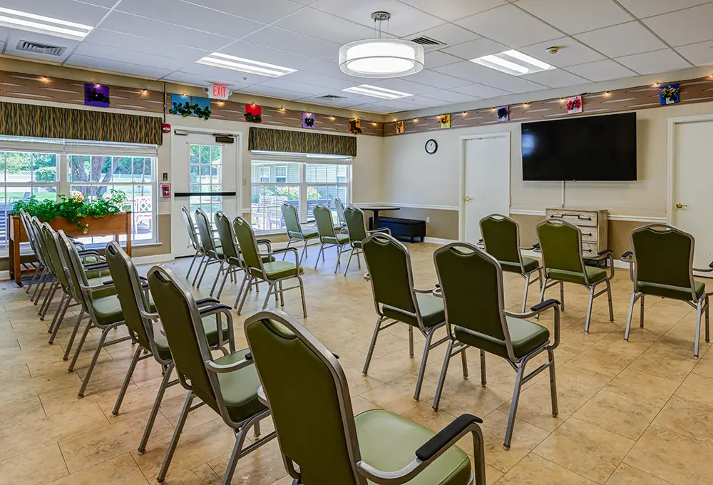 WAR_9273_4_5_6_7 Room with arranged chairs facing a large TV screen, bright lighting, tiled floor, and windows along the wall; decorations and a clock are visible.