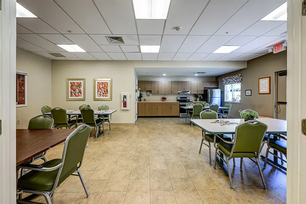 WAR_9283_4_5_6_7 A communal kitchen and dining area with green chairs, tables, tiled floor, wall art, and a kitchenette with stainless steel appliances in the background.