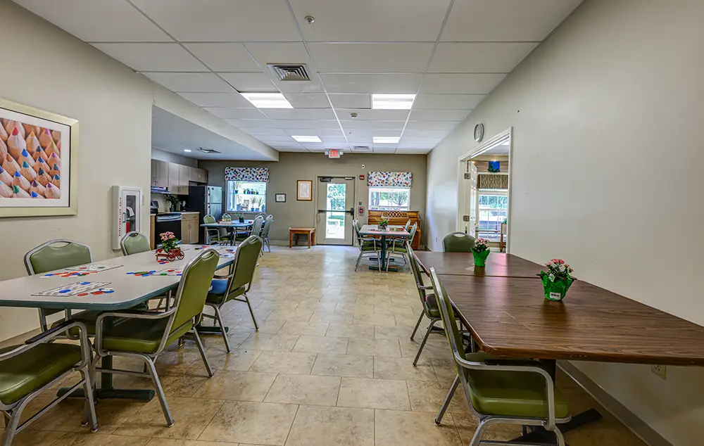 WAR_9293_4_5_6_7 A clean, well-lit common room with tables, chairs, small potted plants, and a visible kitchen area in the background.