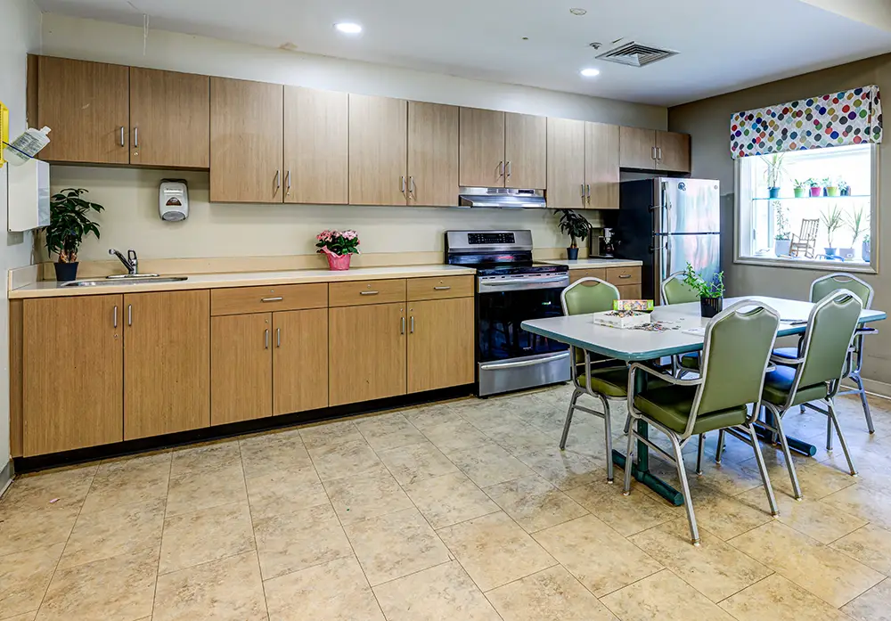 WAR_9303_4_5_6_7 A clean kitchen with wood cabinets, stainless steel appliances, a table with green chairs, potted plants, and a window with a colorful valance.