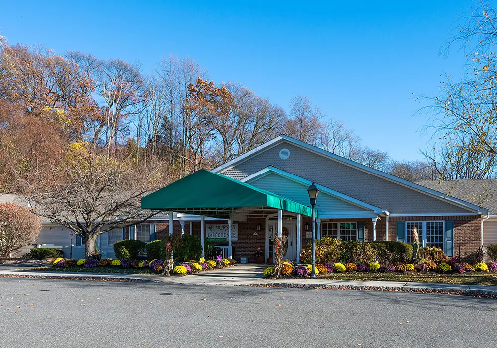 Single-story brick building with a green canopy at the entrance, Arden Courts of Wayne offers memory care, surrounded by shrubs and autumn trees under a clear blue sky.