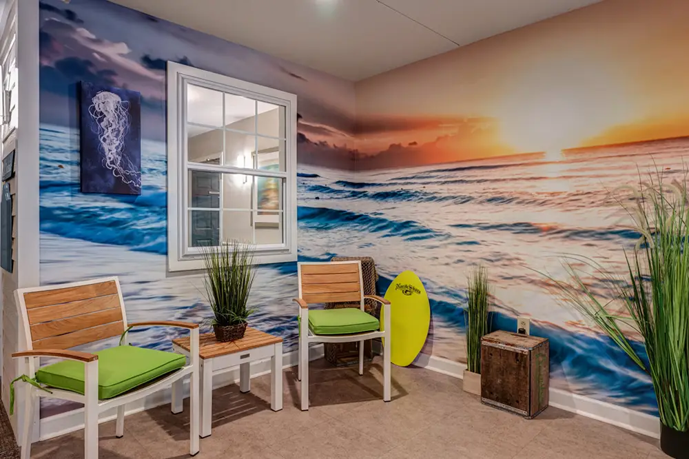 Room with two wooden chairs, a small table, potted plants, a surfboard, and a wall mural of an ocean sunset; window reflects interior lights.