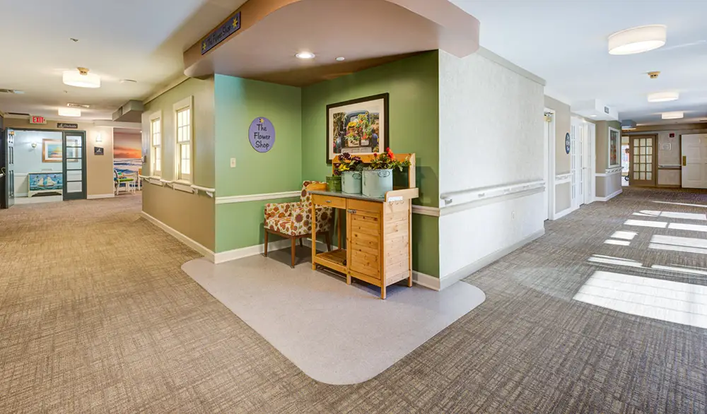 A hallway in a building with a small floral shop nook, containing a chair, a wooden table with flowers, and framed art on a green accent wall.