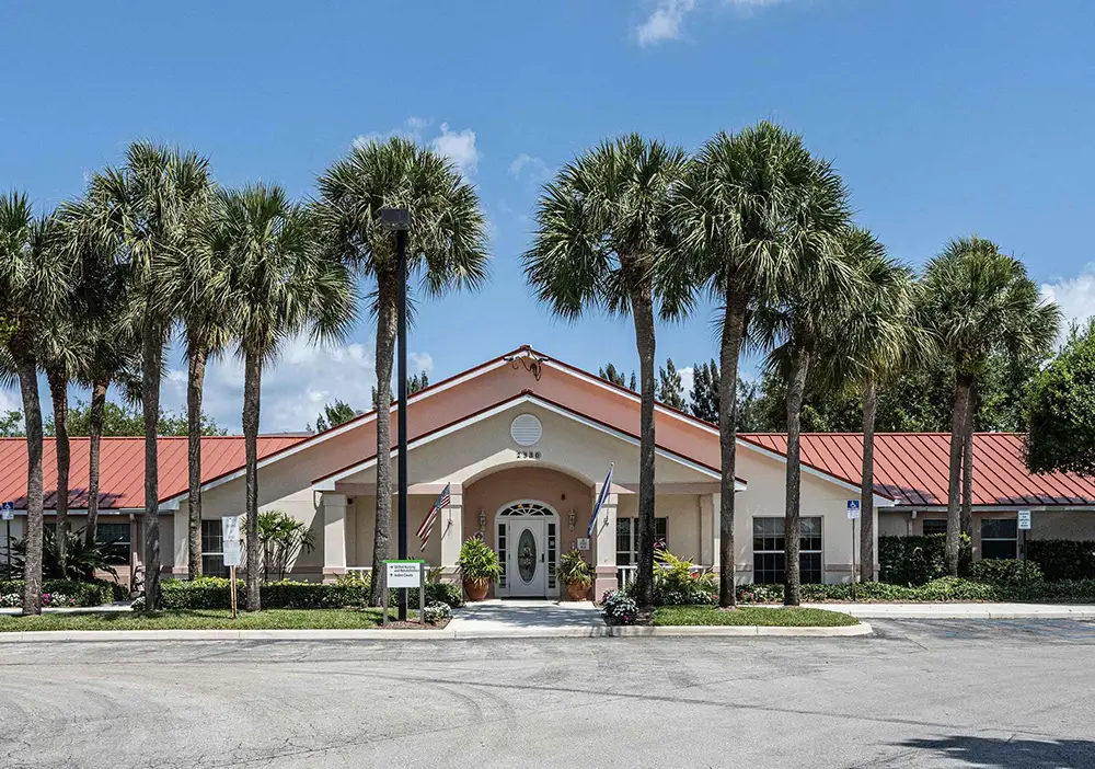 Single-story building with a red roof, central entrance, and several tall palm trees in front—this Arden Courts memory care community is set against a blue sky with scattered clouds in West Palm Beach.