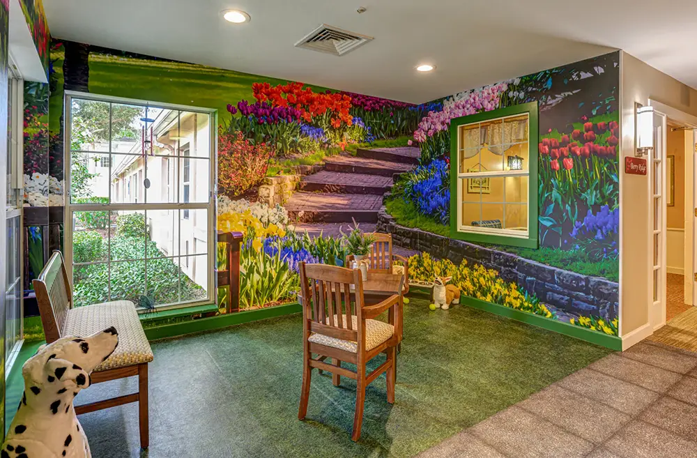 A small seating area with a table and chairs is set beside a large window and mural of a colorful garden with flowers and stone steps. A Dalmatian dog statue sits on a bench.