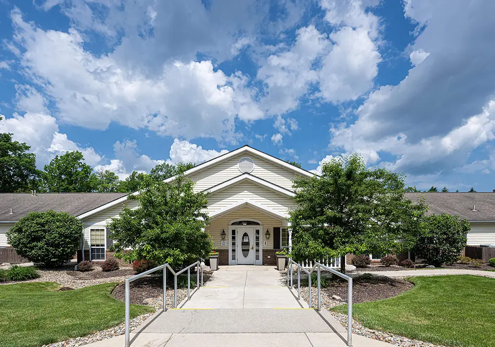 A single-story building with a gabled roof, white siding, and a central entrance, Arden Courts of Westlake is surrounded by landscaped bushes and trees under a partly cloudy sky—designed to offer specialized memory care.