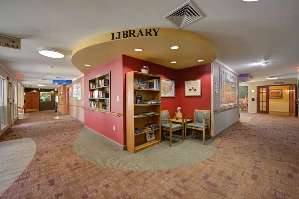 A small library nook with bookshelves, two chairs, and a table is situated in a hallway with brown carpet and red accent walls.