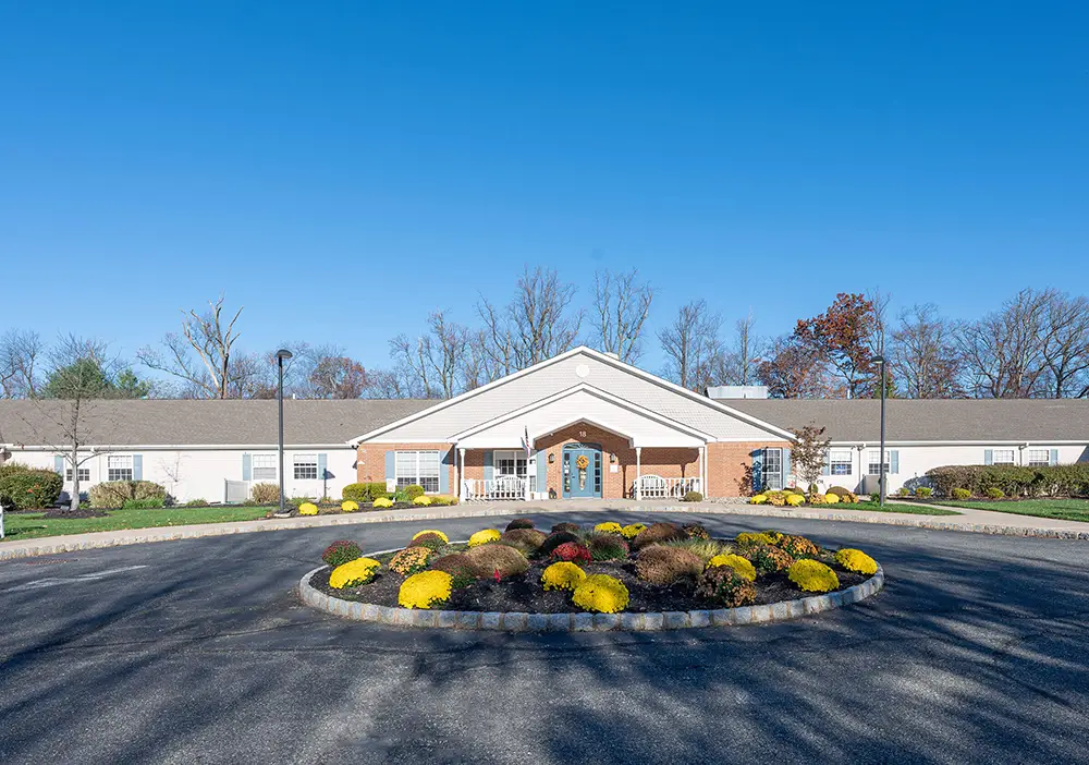 Single-story Arden Courts building in Whippany with a brick entrance, white siding, and a circular driveway featuring landscaped yellow and red shrubs—designed for comfort and specialized memory care under a clear blue sky.