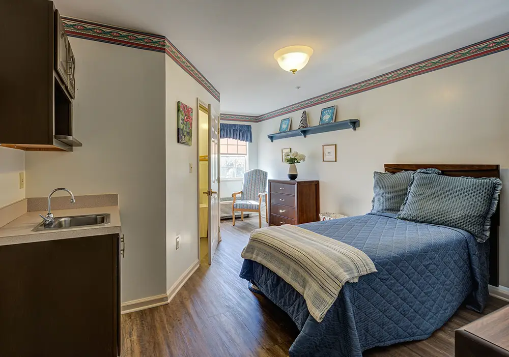 A small, neatly arranged bedroom with a single bed, dresser, armchair, and kitchenette; natural light comes through a window at the far end. A small, neatly arranged bedroom with a single bed, dresser, armchair, and kitchenette; natural light comes through a window at the far end.