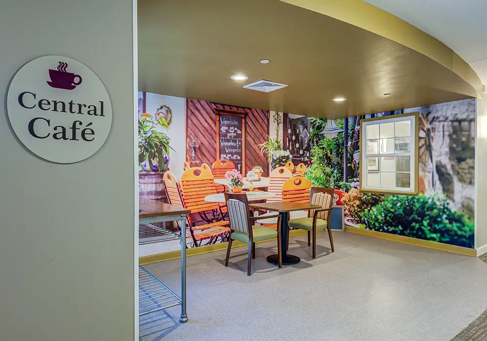 A small seating area with colorful chairs and a mural of an outdoor café scene, located next to a sign reading “Central Café. A small seating area with colorful chairs and a mural of an outdoor café scene, located next to a sign reading "Central Café.